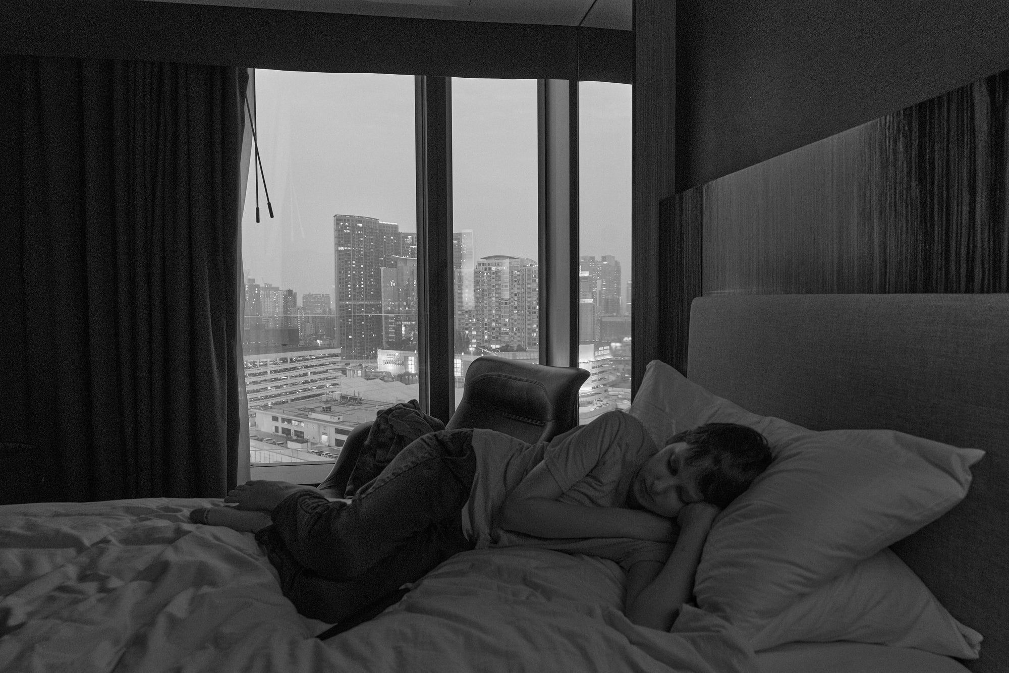 A black and white photograph depicting a monochromatic scene with the Hong Kong city skyline visible through a window, contrasting with a indoor setting.