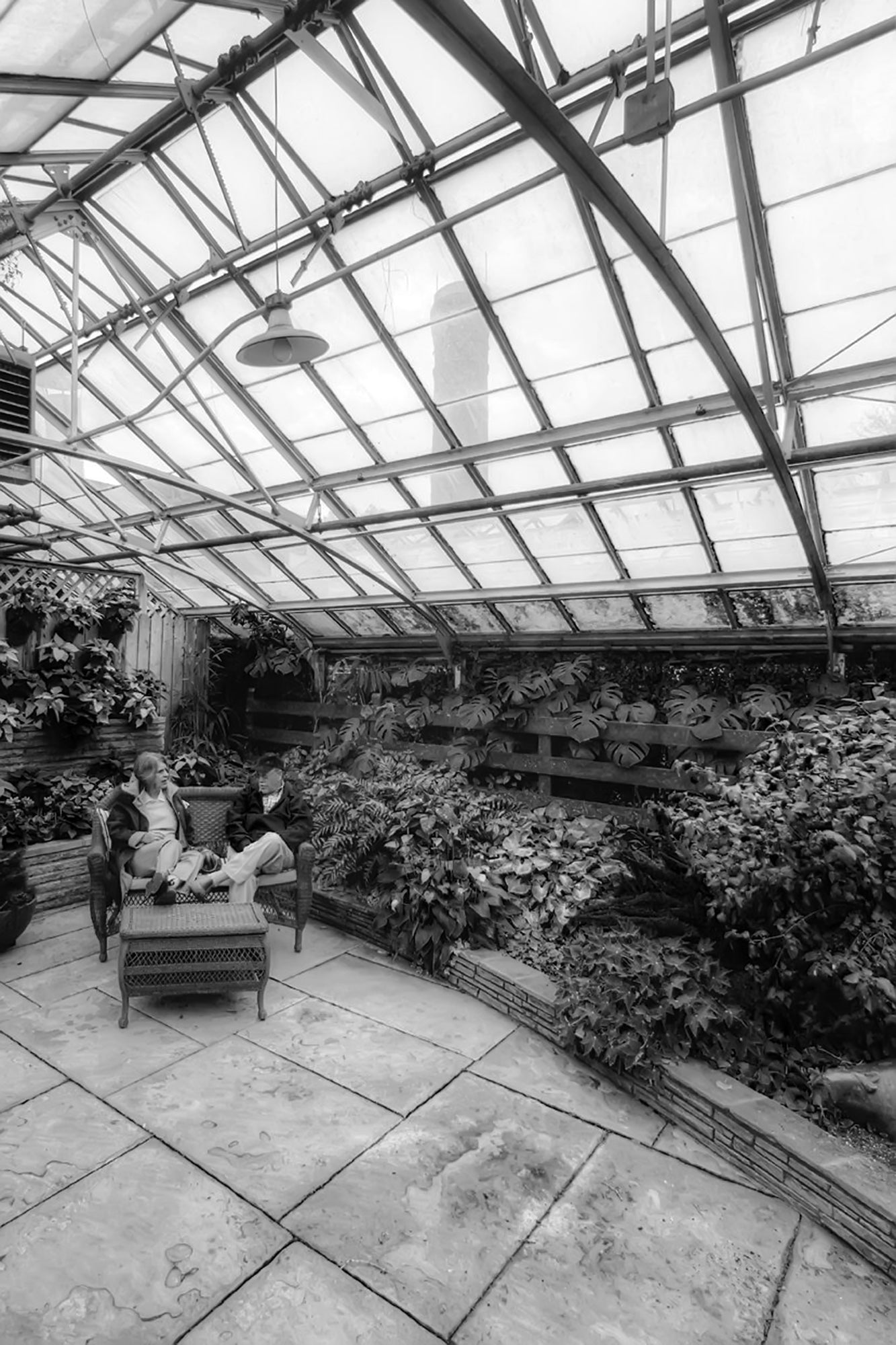 Two people seated on a bench under a greenhouse-style roof, with plants inside and a clear glass structure above.