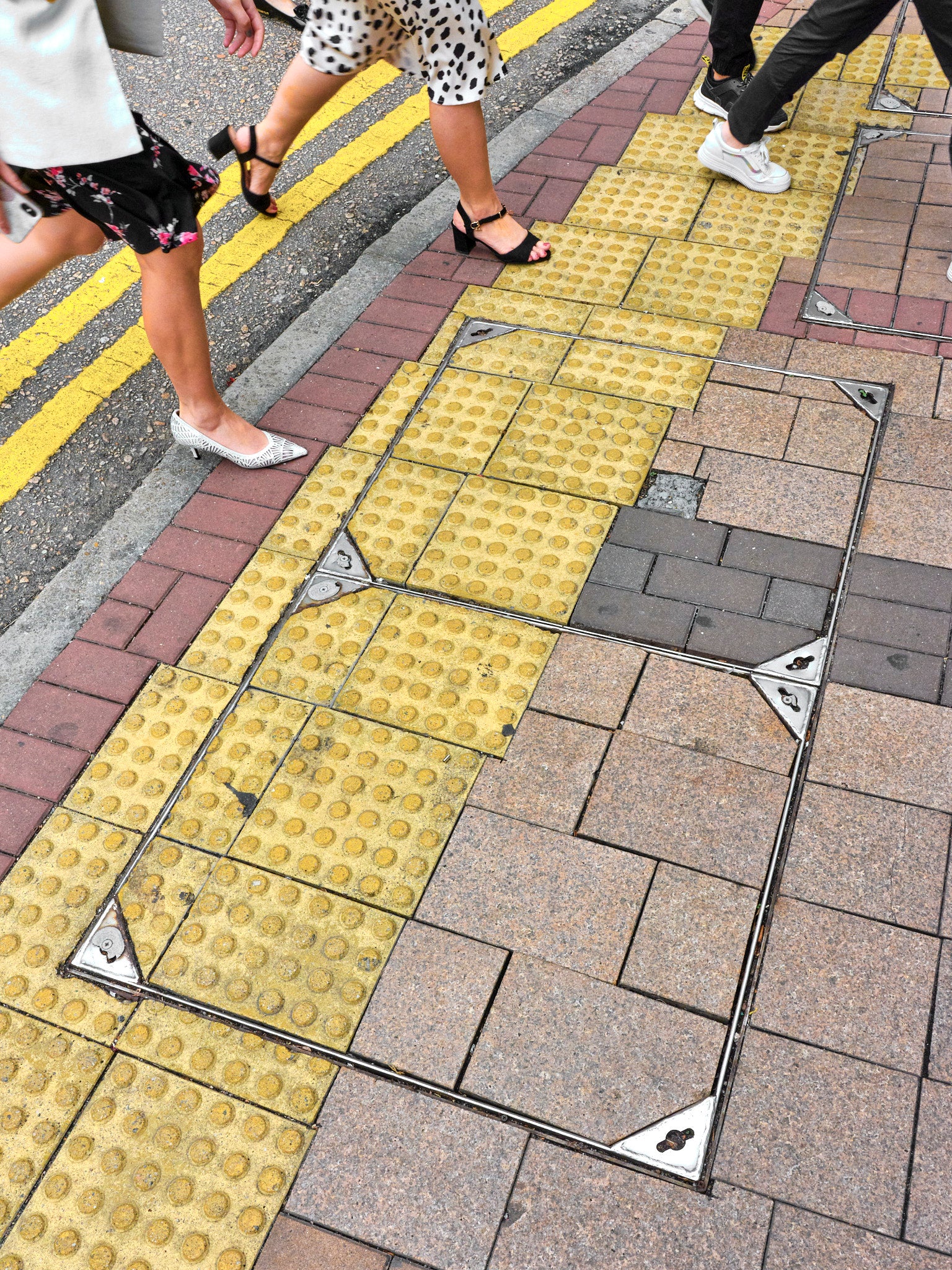 A close-up of a yellow tactile paving on a pedestrian walkway with metallic fixtures, surrounded by regular brick paving, with people walking in the background.