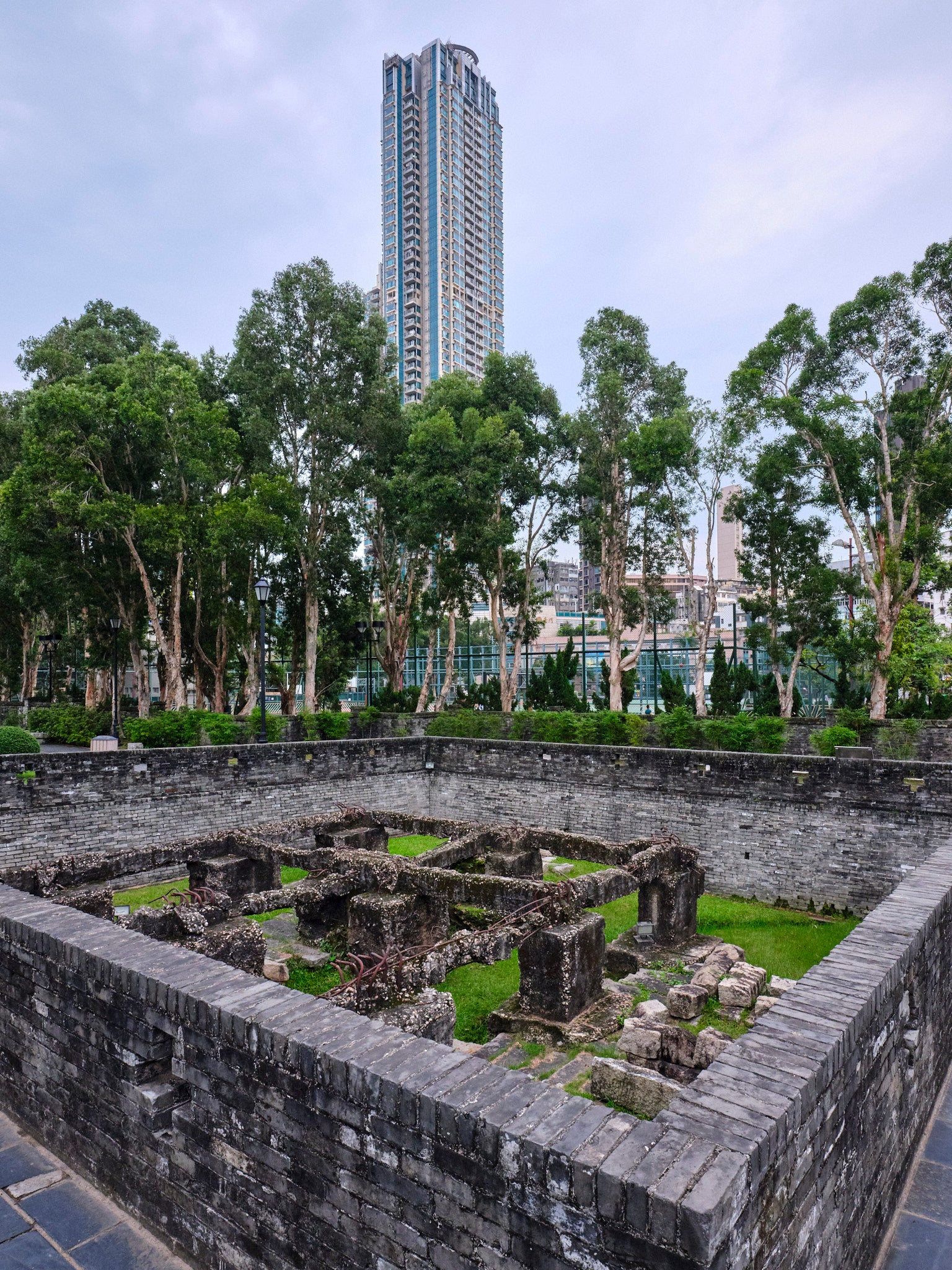 An old ruinous structure of the Hong Kong Walled City, set against a modern high-rise building and a backdrop of trees and sky.