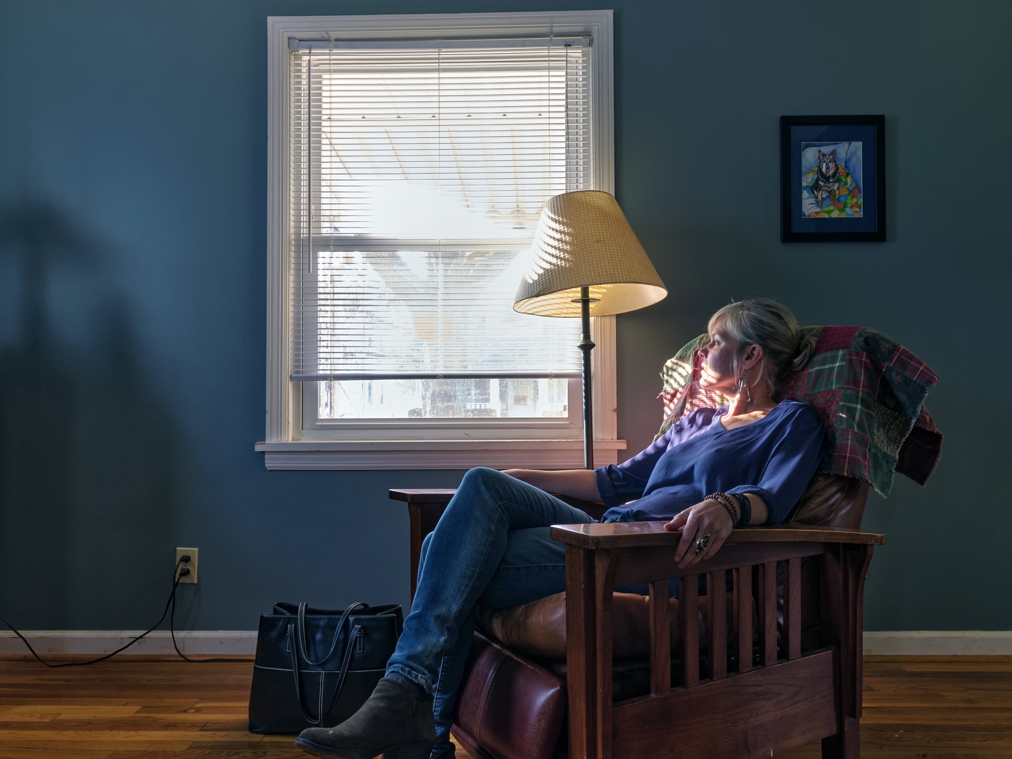 A person seated in a chair near a window, with a lamp and a dog painting in the background, captured in natural light.