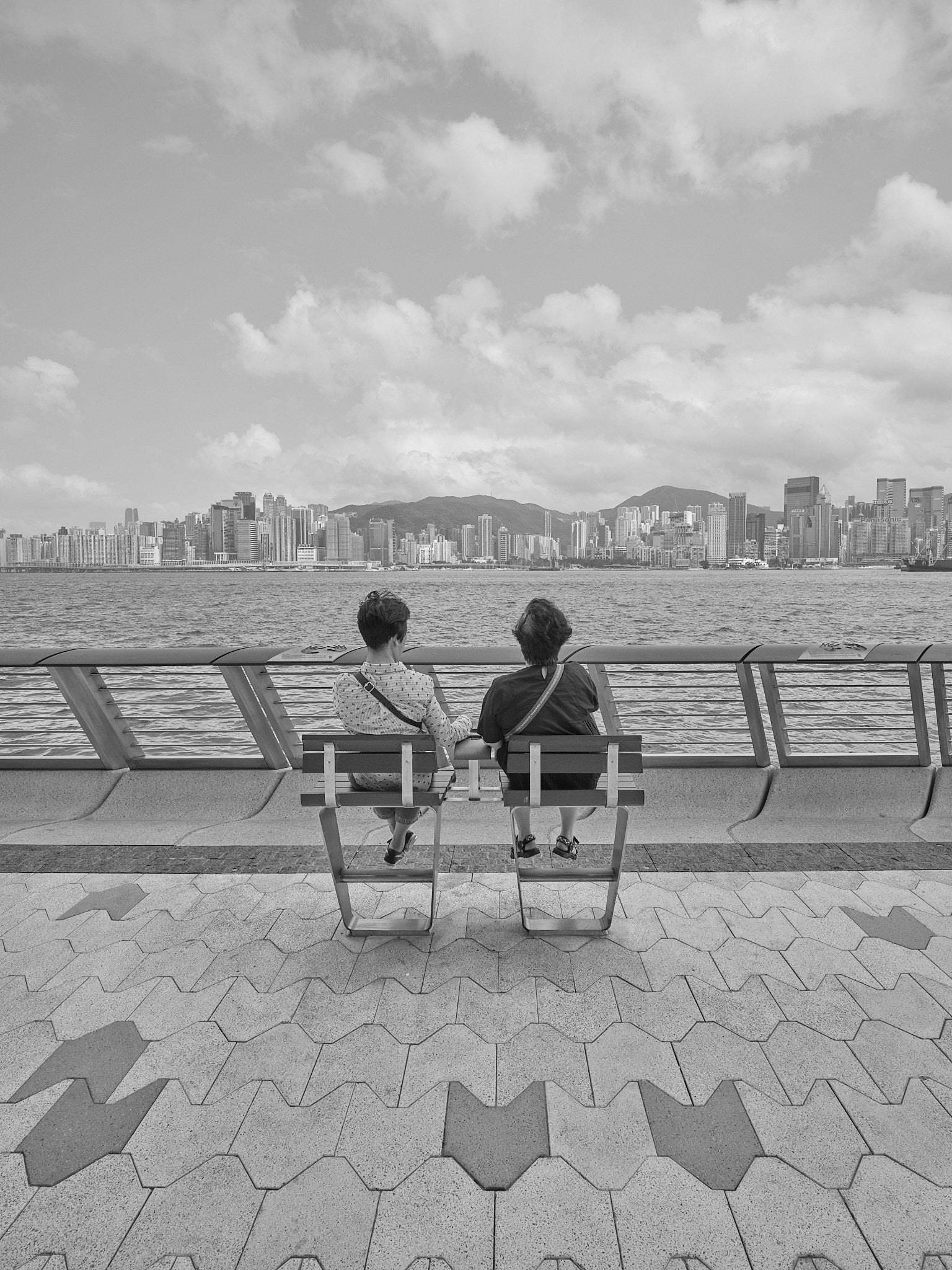 Two people sitting on benches in front of Victoria Harbor in Hong Kong with the city skyline in the background, in black and white.