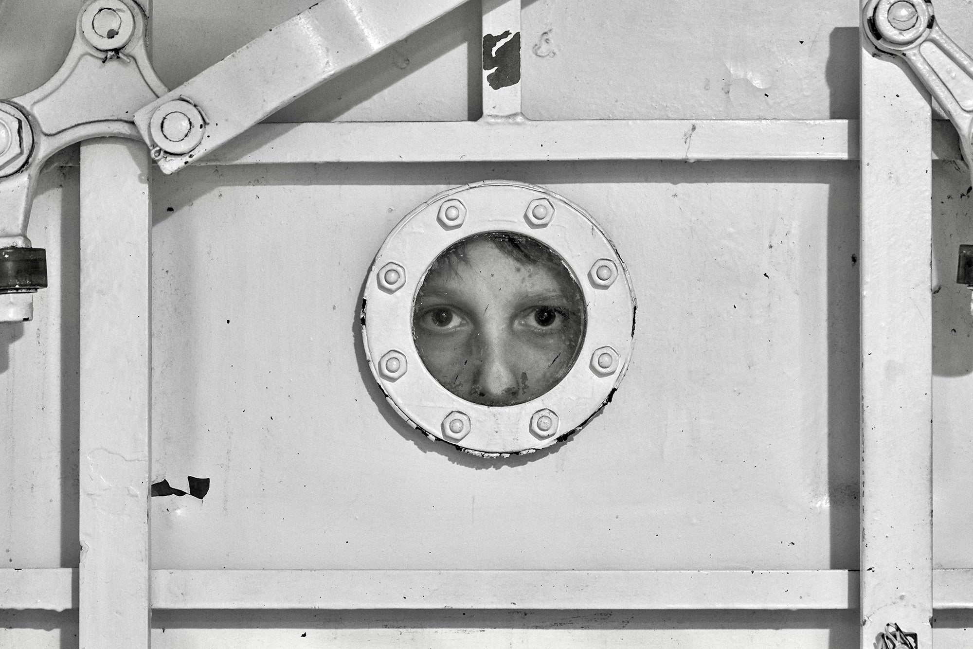 A black and white photo of a person's face looking through a viewfinder or peek hole on a secured metal door.