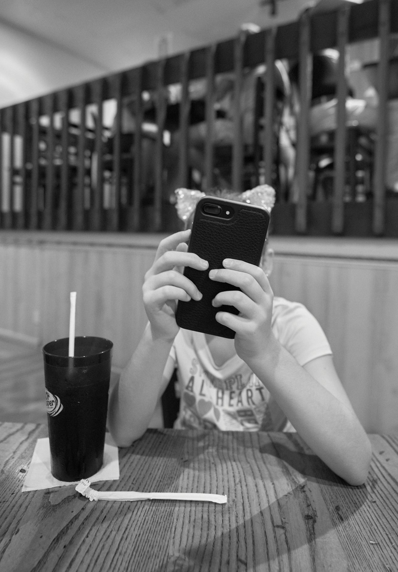 A black and white photo of a person sitting at a table with a drink, holding a smartphone up to their face.