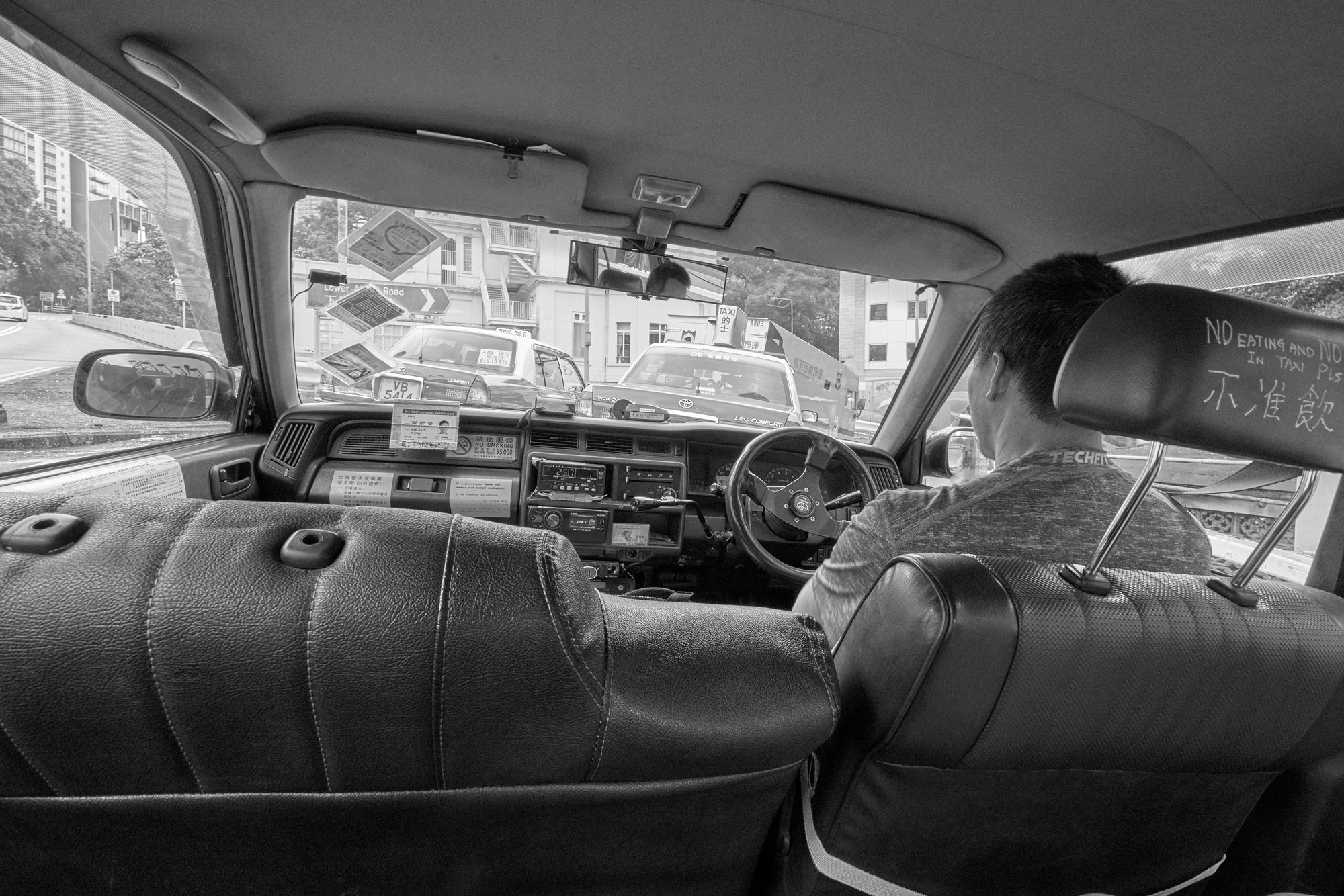 A black-and-white photograph of the interior of a taxi in Hong Kong's Mid-Levels, showing a driver at the wheel with a dashboard covered in signs and a cityscape visible through the windshield.