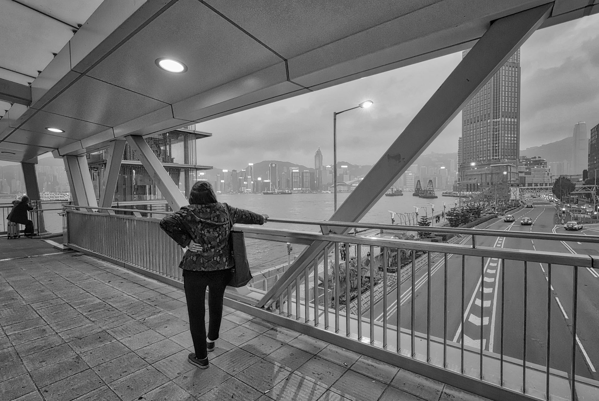 Black and white photograph of a person standing on a bridge with a view of Hong Kong's skyline and Victoria Harbor in the background.