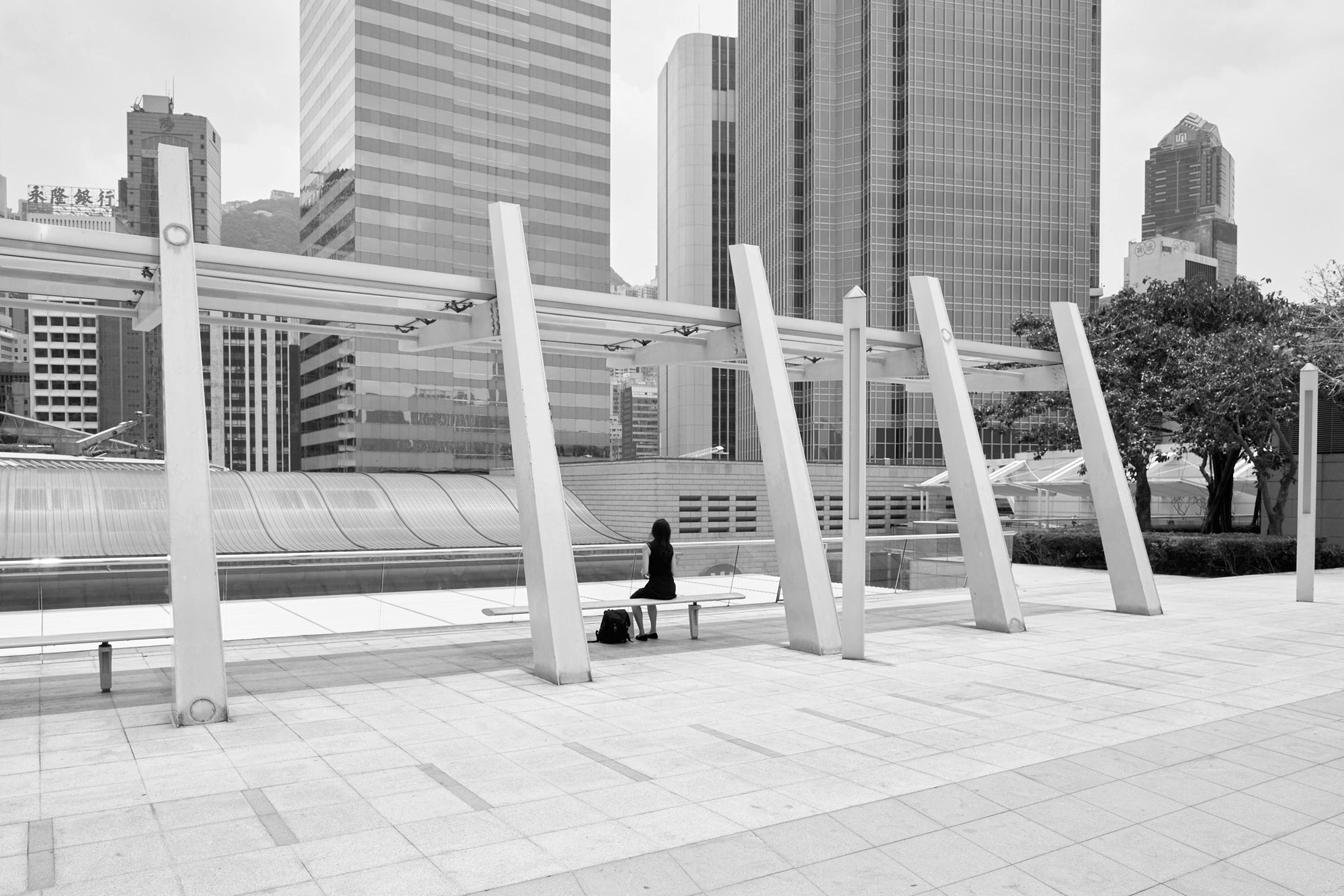 Black and white photograph of a person sitting on a bench in Hong Kong with tall buildings in the background and artistic structures in the foreground.