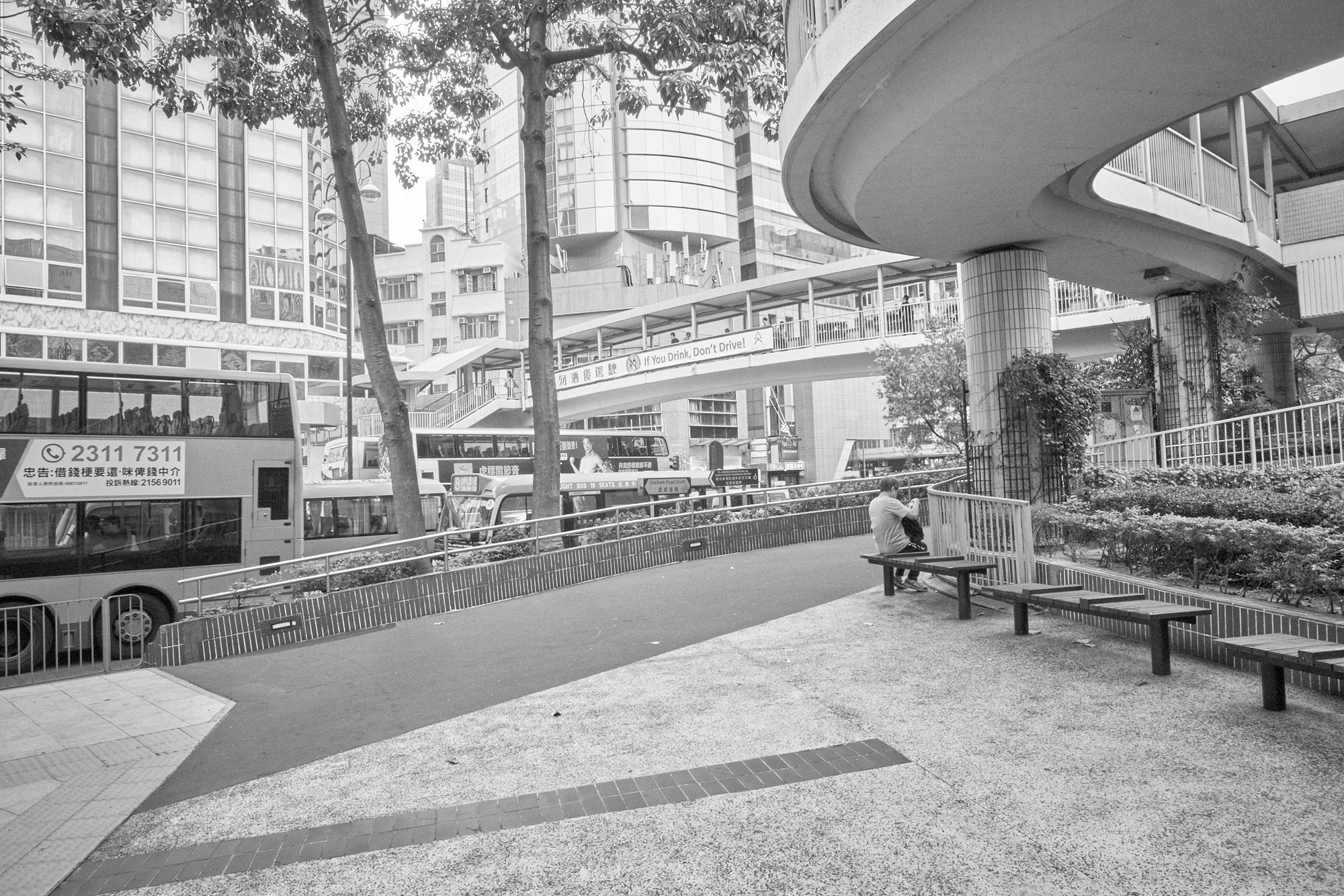 Black and white photograph of an urban scene in Hong Kong featuring a skyline, streets, and a person sitting on a bench.