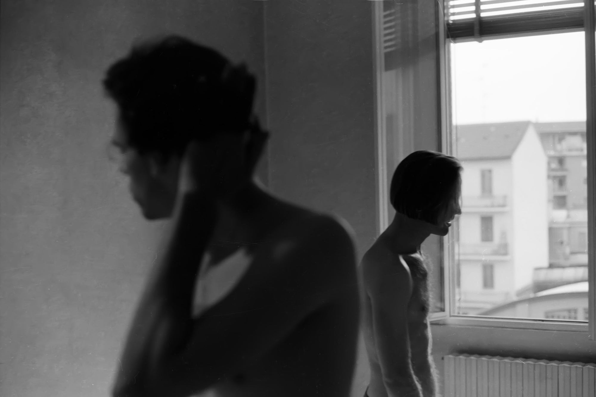 A black and white photo showing two people from the side, standing near a window with an outdoor view.
