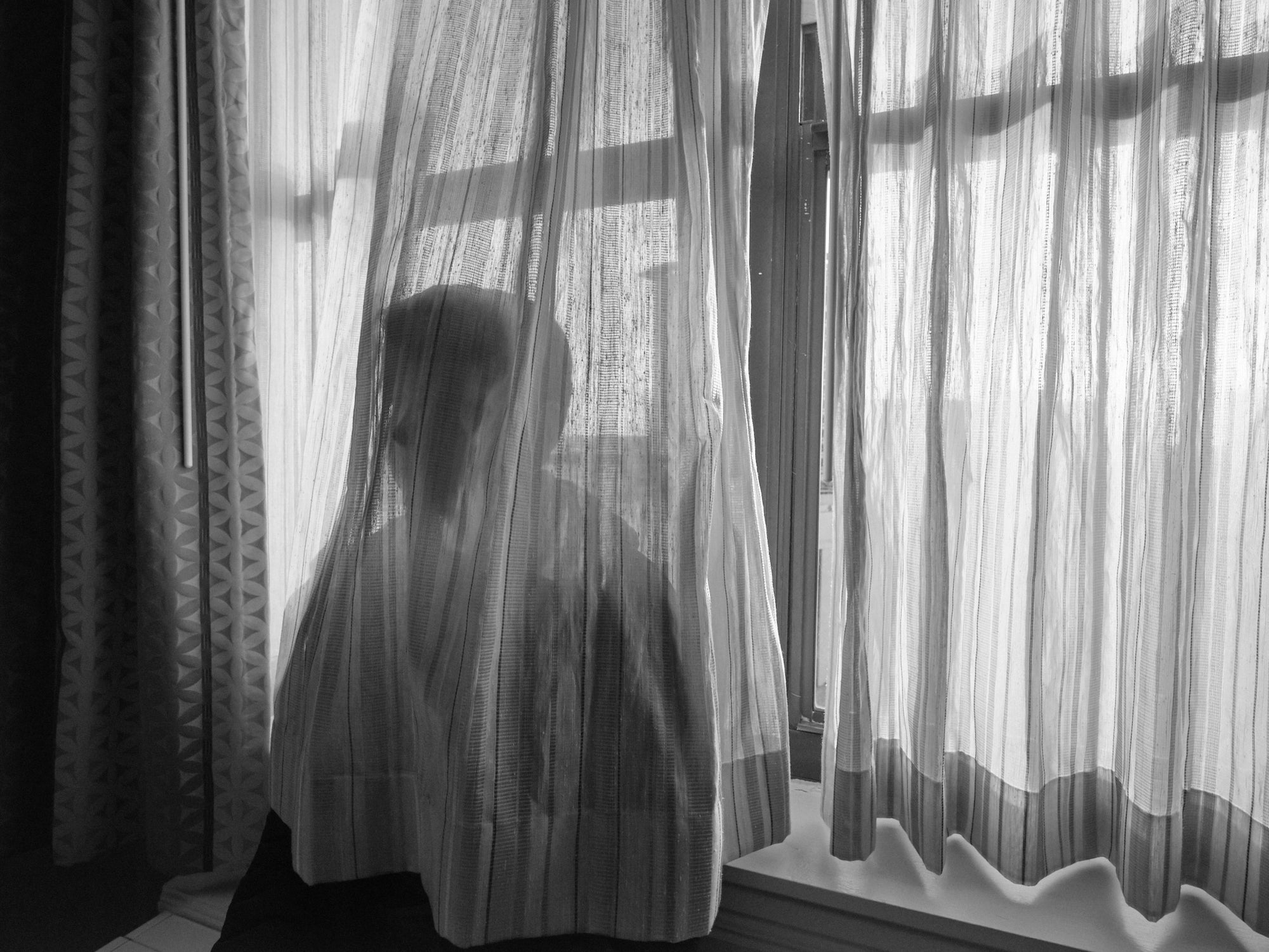 A black and white image of a person standing behind a window with a white curtain, partially obscured by the curtain.